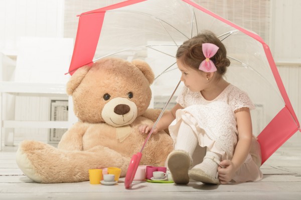 niña pequeña sentada bajo un paraguas con su oso de peluche