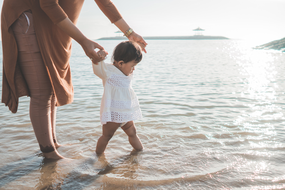 mamá sosteniendo las manos de su bebé que camina en la playa